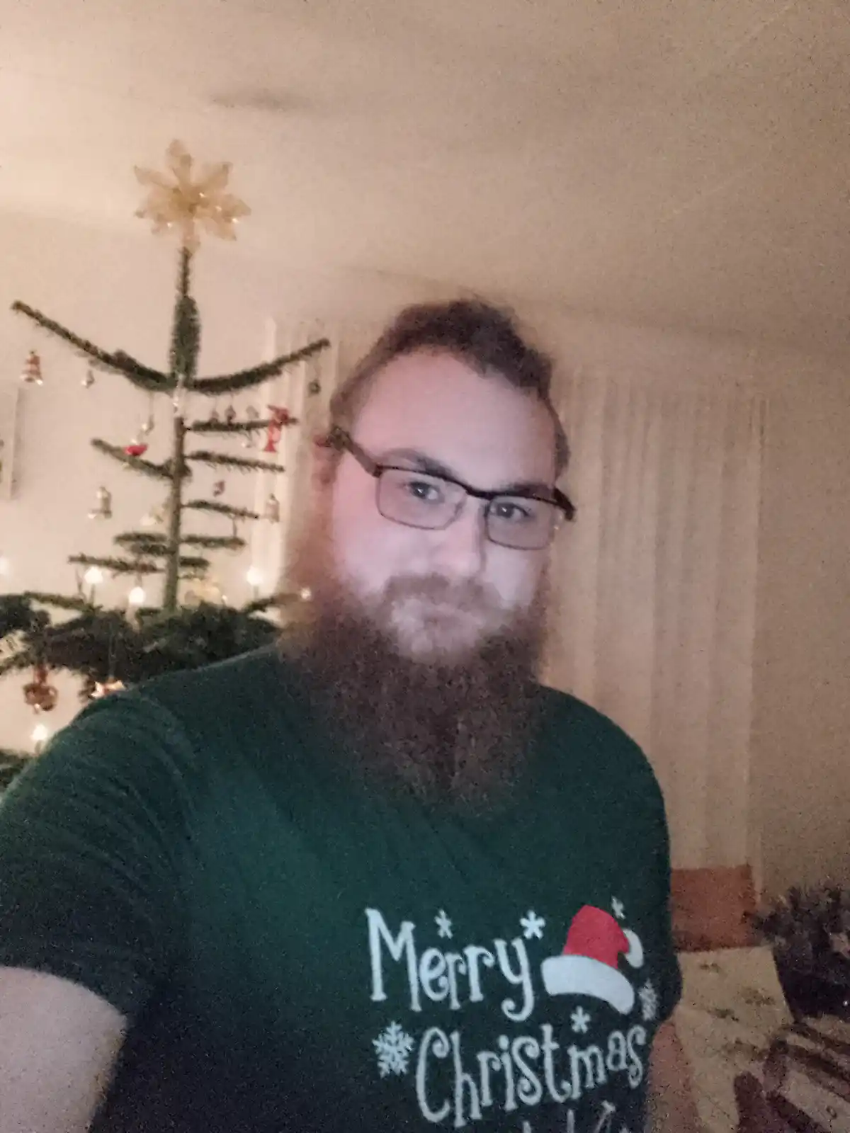 A man with a full beard and glasses wears a green t-shirt with 'Merry Christmas' and a Santa hat graphic, standing in front…