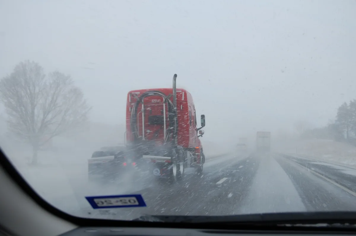 A view from inside a car during a heavy snowstorm, showing a red semi-truck ahead on a highway. Snowflakes obscure the…