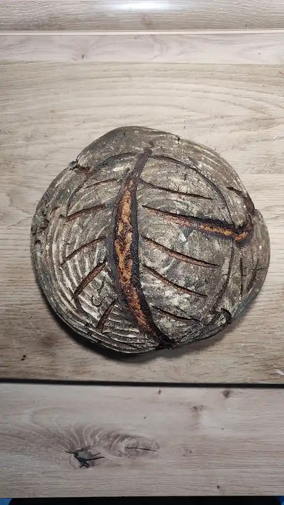 A round, rustic loaf of bread with a dark, cracked crust and deep scoring lines sits on a light wood surface. The scoring…