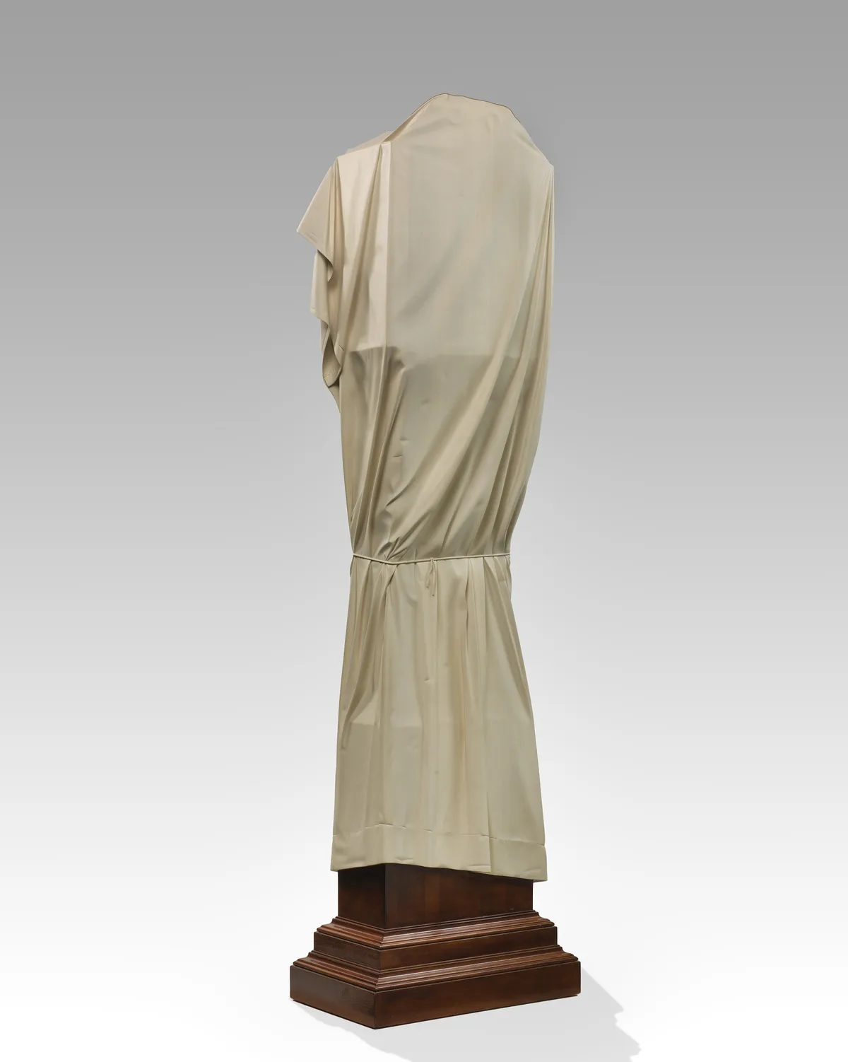 A full-length statue, likely classical in style, is completely covered from head to toe in a beige, draped fabric, obscuring…