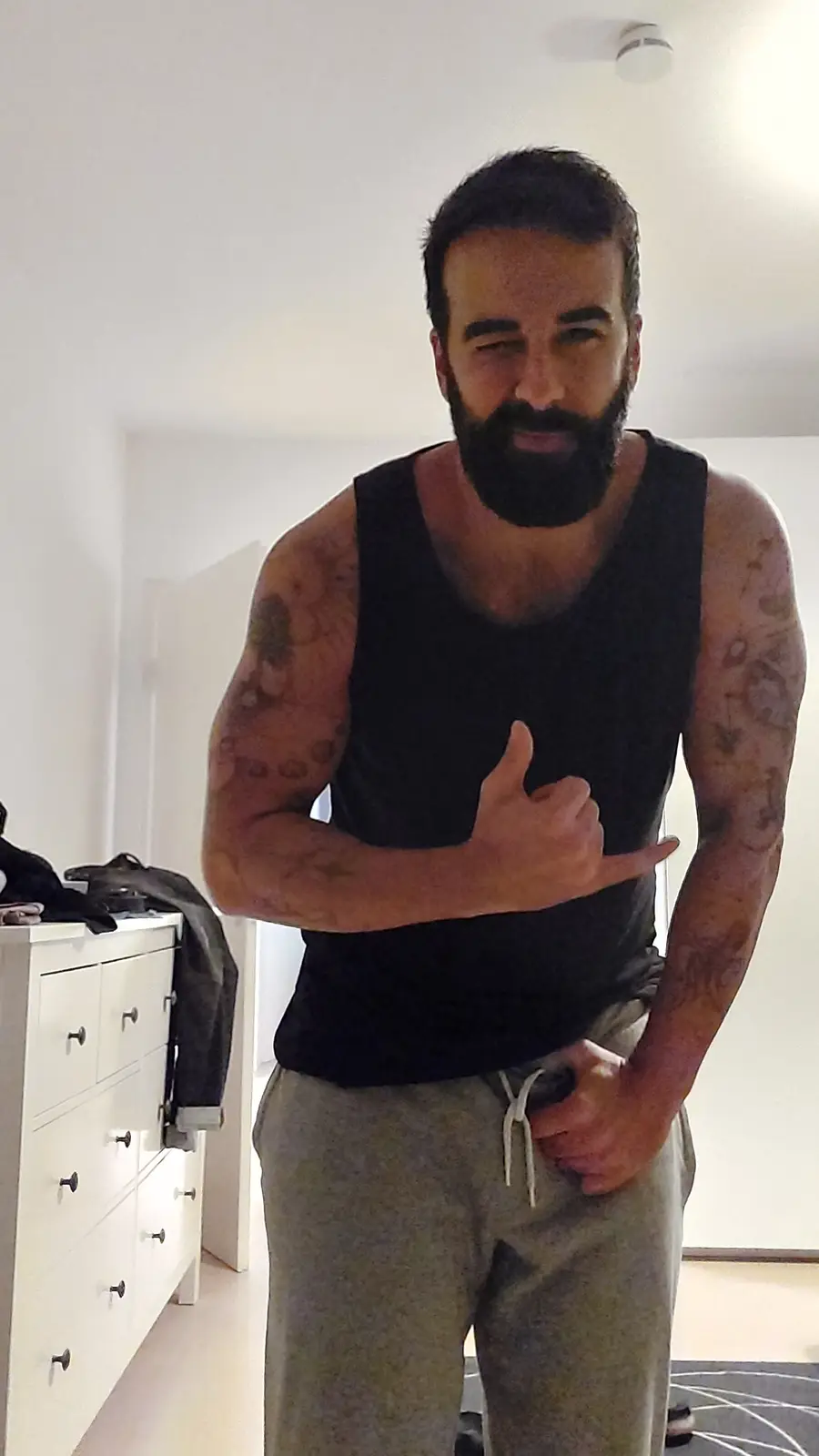 A man with a full beard and tattoos covering both arms stands in a room wearing a black tank top and grey sweatpants. He…