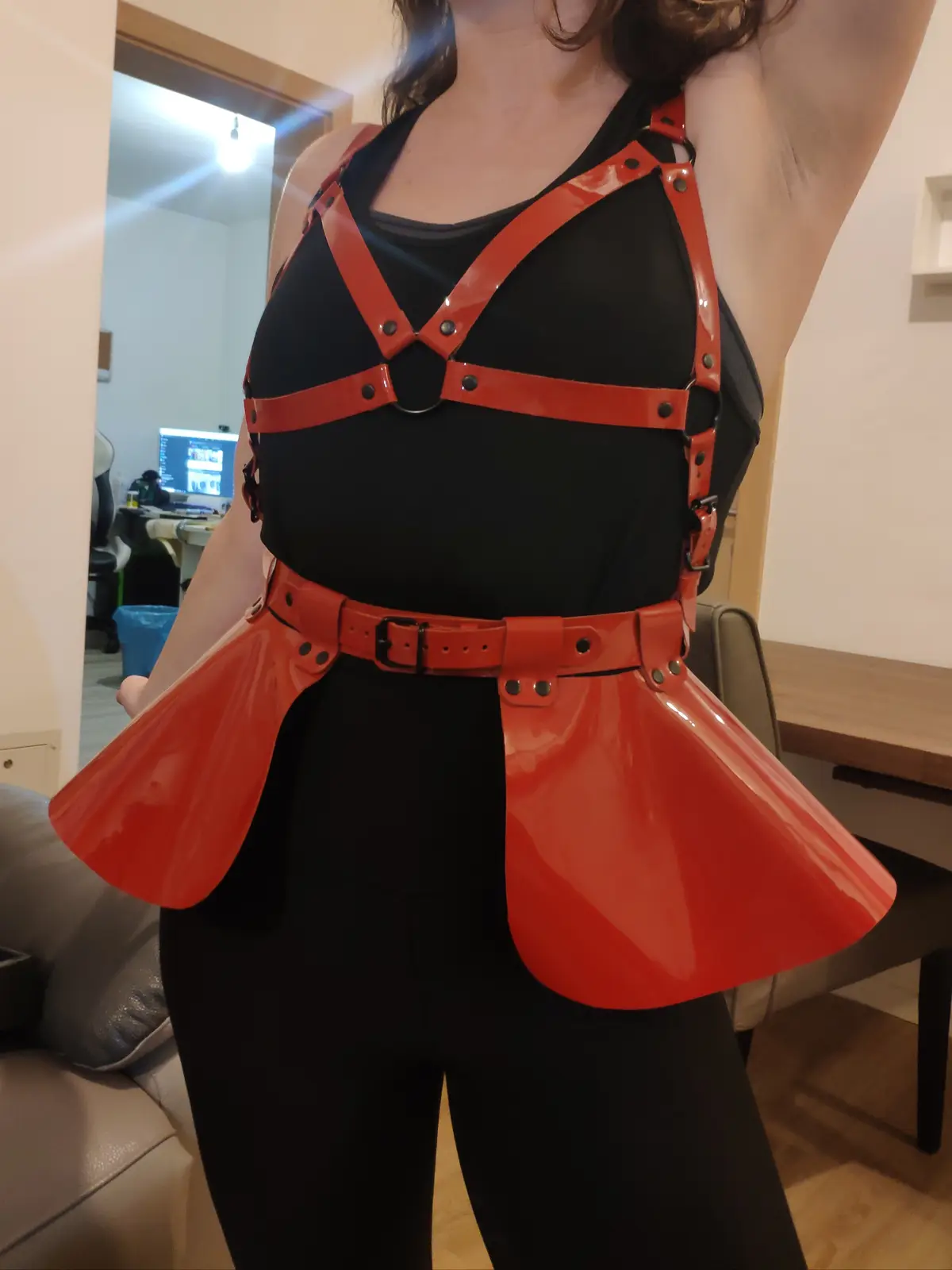A person wearing a black tank top and leggings is shown from the chest down, adorned with a bright red, glossy, harness-like…