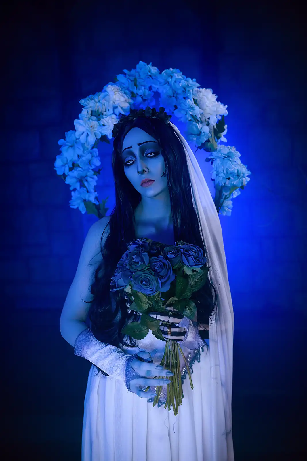 A person dressed as a bride with pale makeup, long dark hair, and a white veil, wearing a floral crown and holding a bouquet…