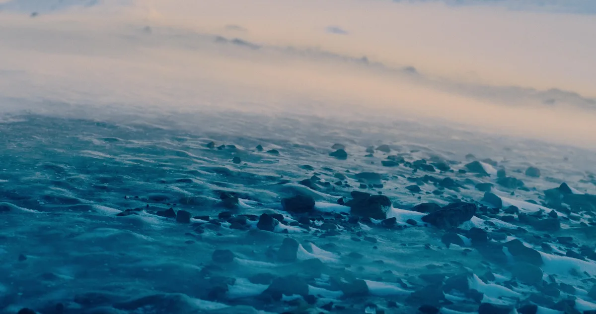 A serene, low-angle photograph of a misty, icy shoreline at dawn or dusk, featuring dark rocks partially covered in snow and…