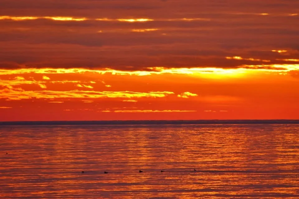 A serene sunset over water with vibrant orange and red hues reflecting on the calm surface, featuring several small dark…