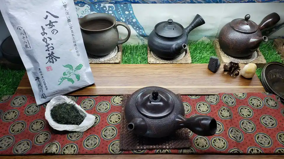 A still life of traditional Japanese tea ceremony items including several ceramic teapots, a bag of green tea labeled…