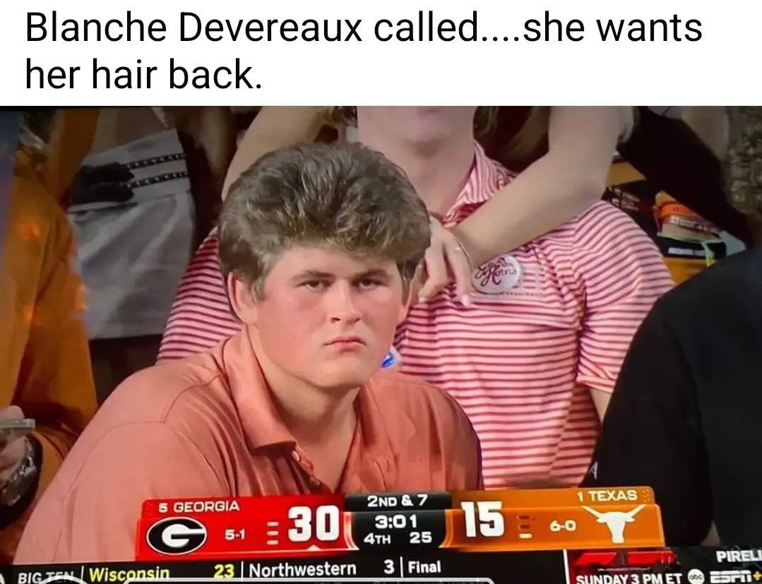 A still image from a college football game shows a young man with a large, curly, light-brown hairstyle looking intensely…