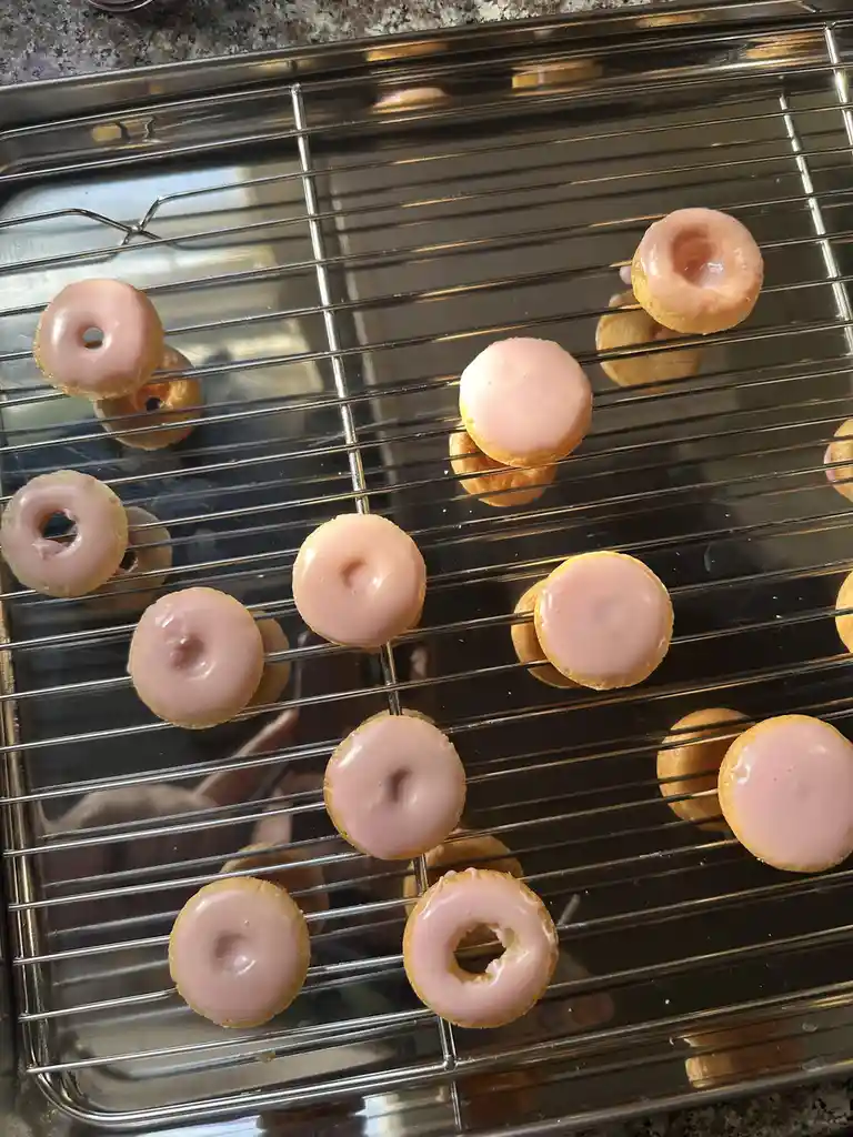 A top-down photo shows numerous small, round, pink-frosted donuts resting on a metal cooling rack. Some donuts are oriented…
