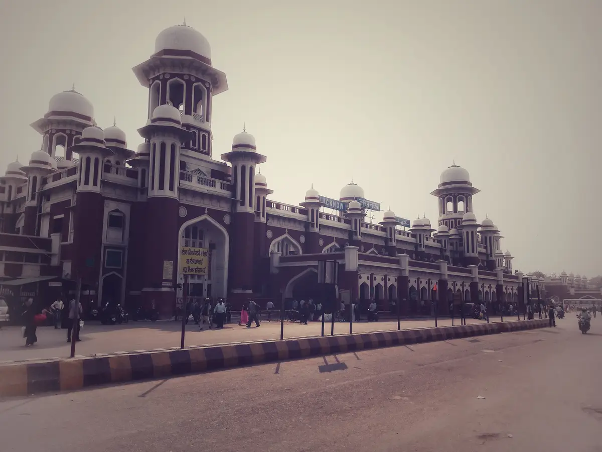 The image shows the exterior of Lucknow Railway Station, a large red and white colonial-style building with multiple domes…