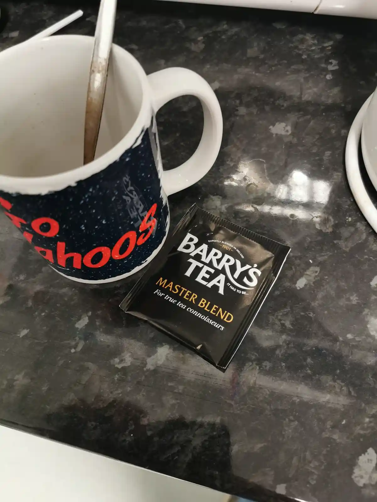 A white ceramic mug with red 'Yahoo!' text and a spoon inside sits next to a black Barry's Tea Master Blend packet on a…