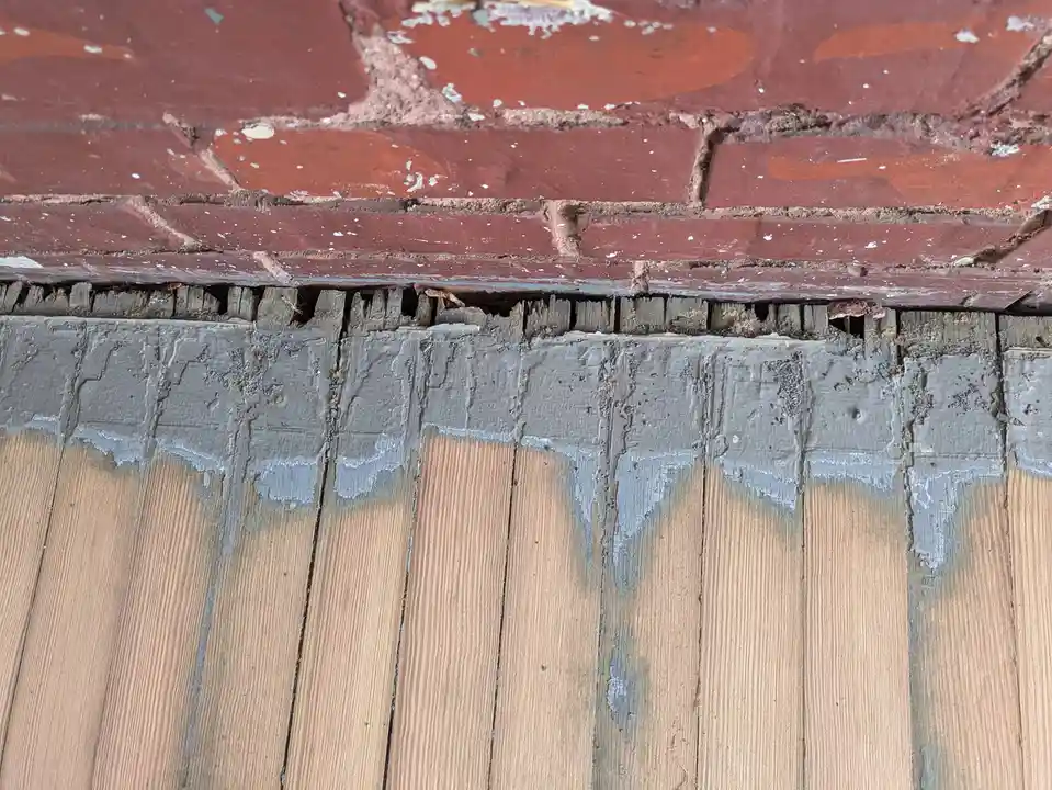 A close-up photo shows a weathered wooden deck with peeling gray paint, situated beneath a red brick wall with visible…
