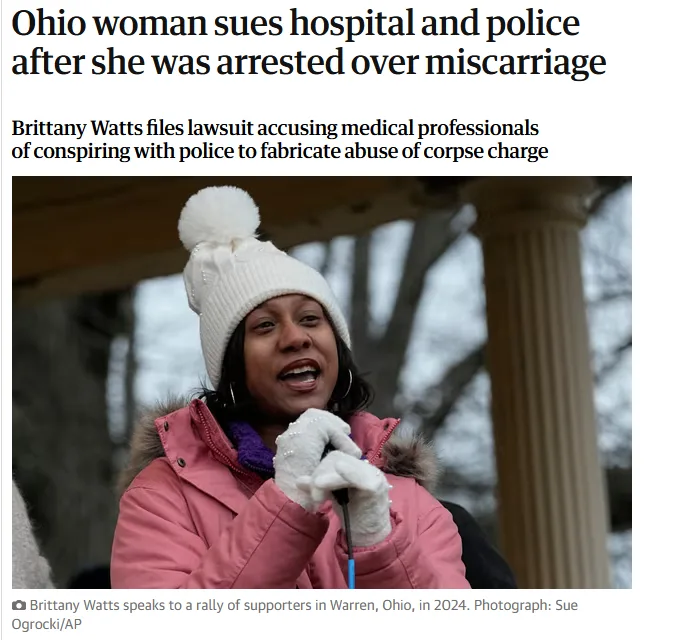 A woman wearing a white knit hat with a pom-pom, a pink winter coat with a fur-lined hood, and white gloves is speaking into…