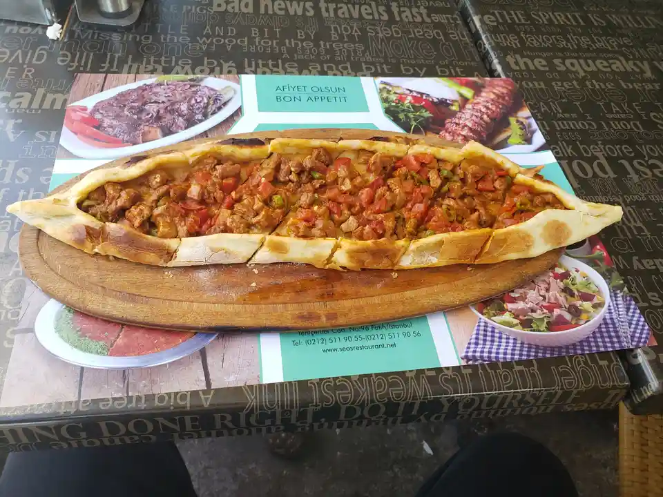 A large, freshly baked Turkish-style meat-filled bread, resembling a pide or lahmacun, sits on a wooden serving board atop a…