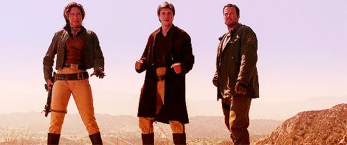 The image shows three men standing side-by-side in a desert landscape, with a large wrecked spaceship behind them. The scene…