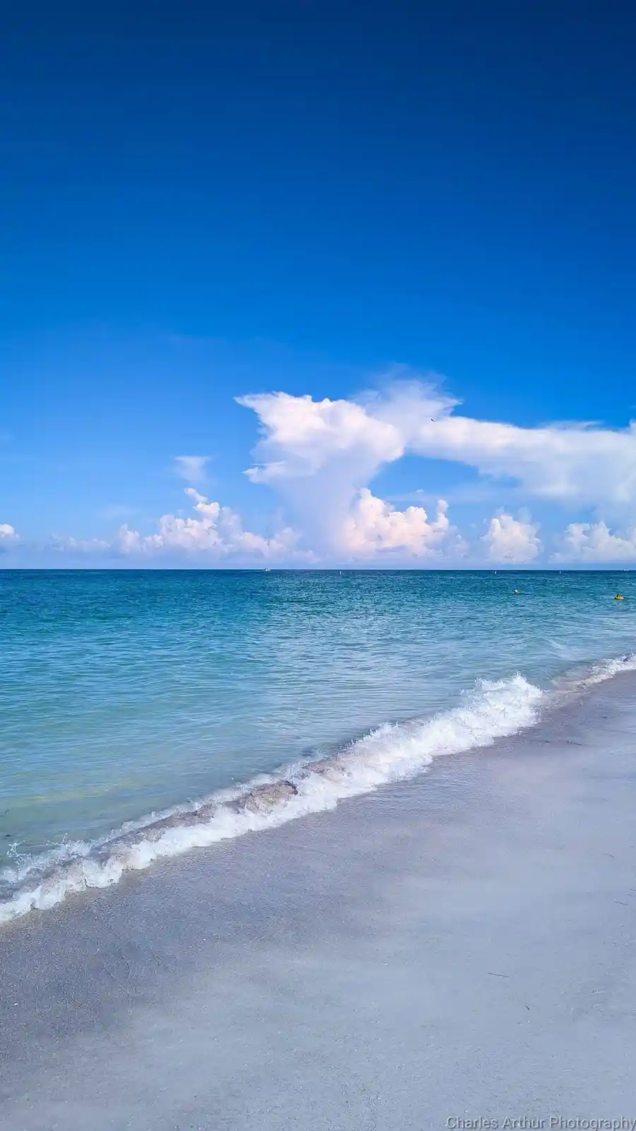 A serene beach scene with turquoise water, white sand, and a large, fluffy cloud formation in a bright blue sky. The…