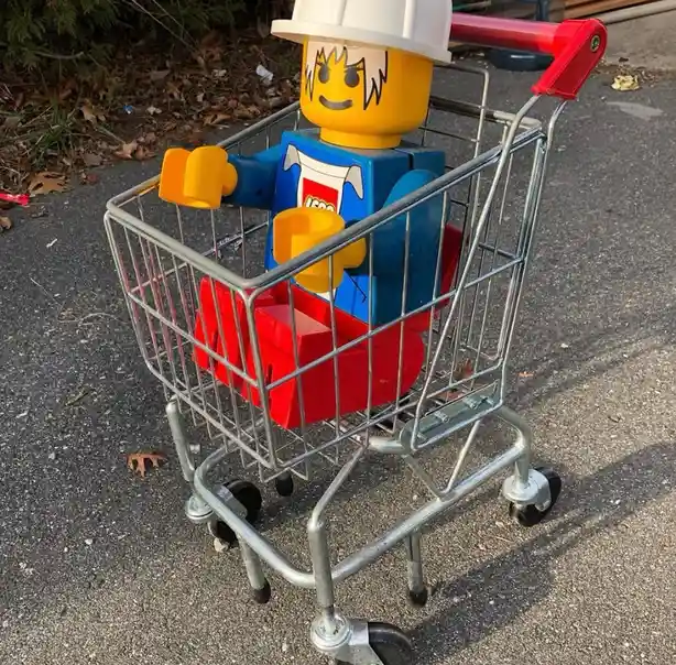 A large LEGO minifigure wearing a white hard hat sits inside a small metal shopping cart on an asphalt surface. The…