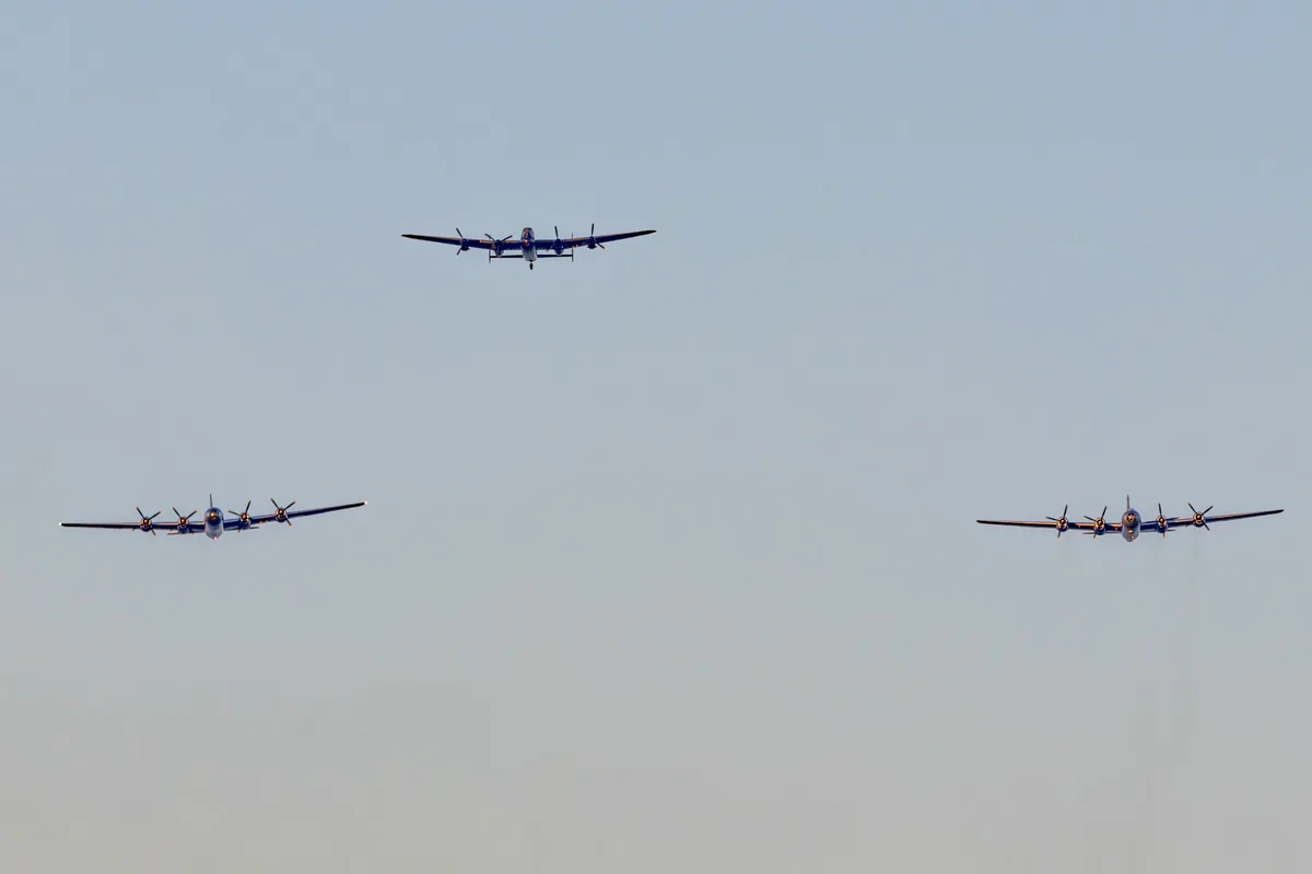 Three vintage-style propeller aircraft fly in formation against a clear sky. The central plane is noticeably smaller and…