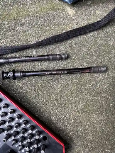 Two black metal rods with engraved specifications lie on a concrete surface next to a red and black socket wrench set. The…