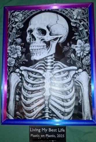 A framed black-and-white artwork of a human skeleton surrounded by floral patterns, with a plaque below reading 'Living My…