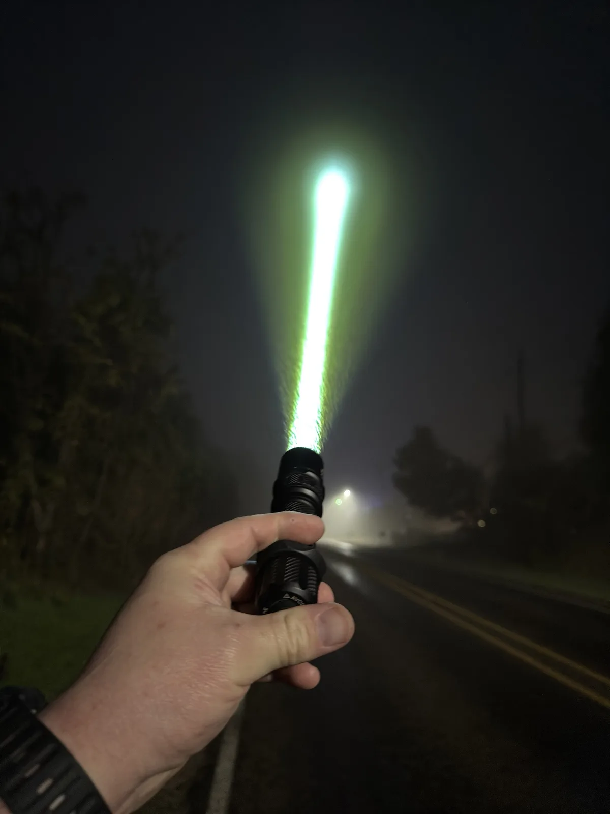 A hand holds a powerful flashlight on a dark, foggy road at night, projecting an intensely bright, narrow green beam upward…
