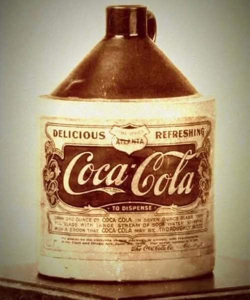 An old, vintage ceramic Coca-Cola dispenser jug with a faded label showing the classic Coca-Cola logo and instructions for…