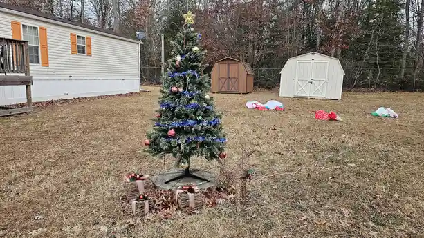 A decorated artificial Christmas tree stands in a dry, brown lawn in front of a house, surrounded by scattered holiday…