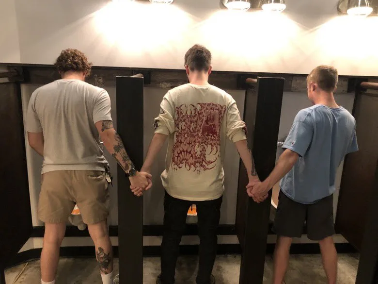 three men standing side by side in a row of public restroom urinals, all facing away from the camera, holding hands with the…