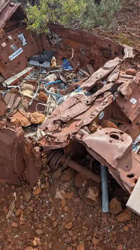A rusted, crushed car wreck lies in a red dirt field, surrounded by rocks and overgrown bushes. Inside the wreckage, a…