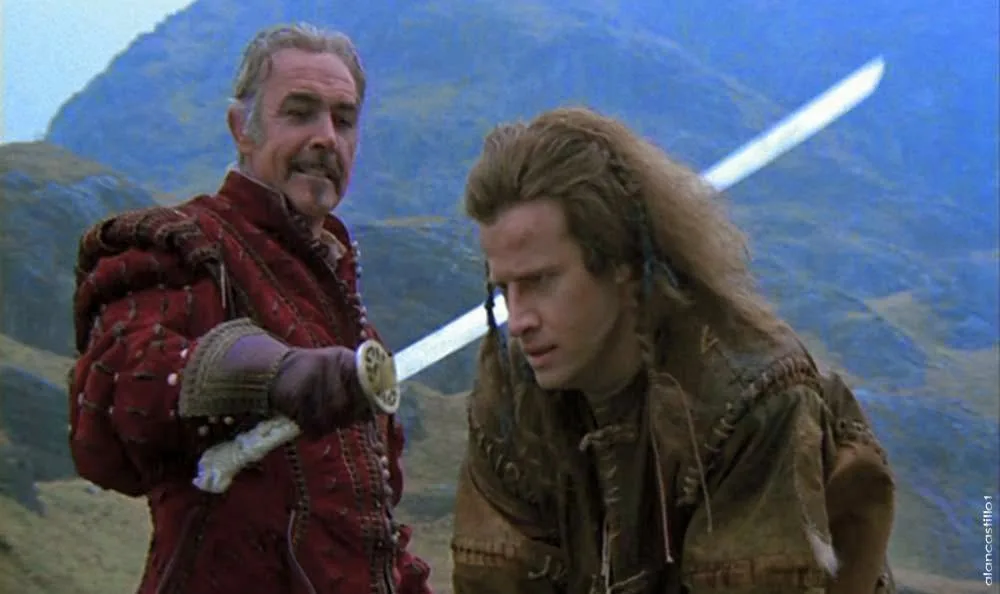 two men in medieval-style clothing stand outdoors with mountains in the background; the man on the left, wearing a red…