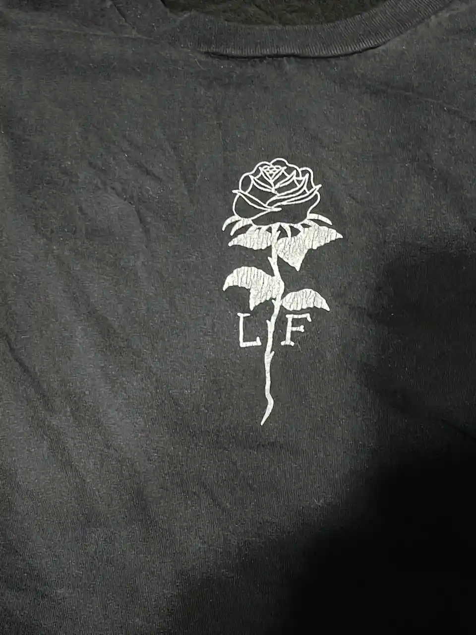 A close-up of a dark gray t-shirt featuring a white outline of a rose with the letters 'LF' printed on its stem. The rose…