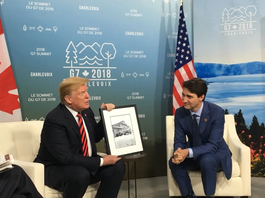 Donald Trump and Justin Trudeau sit in chairs at the 2018 G7 Summit in Charlevoix, Canada. Trump holds up a framed…
