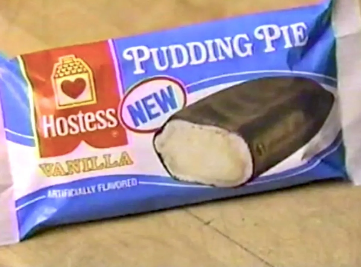 A close-up of a Hostess Pudding Pie snack package, featuring a chocolate-coated vanilla pudding filling, with 'NEW' and…