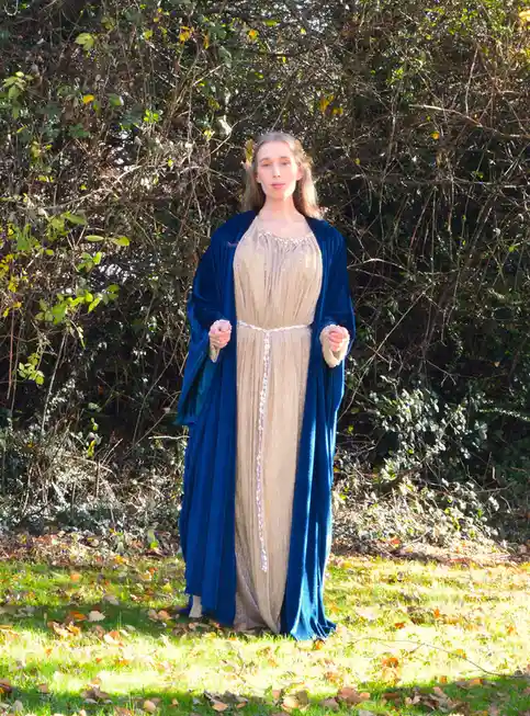 A woman stands outdoors in a grassy area with fallen leaves, wearing an elaborate fantasy costume consisting of a shimmering…
