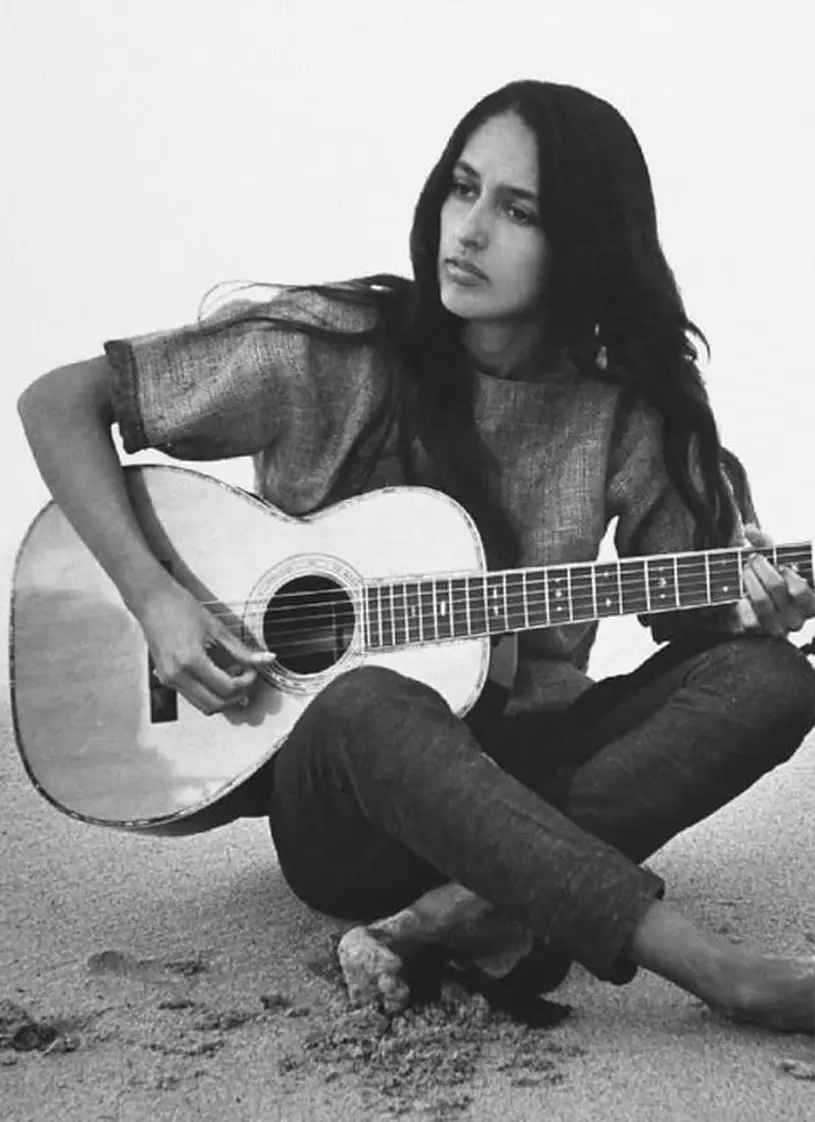 A black-and-white photo shows a woman with long dark hair sitting cross-legged on sand, holding an acoustic guitar. She…