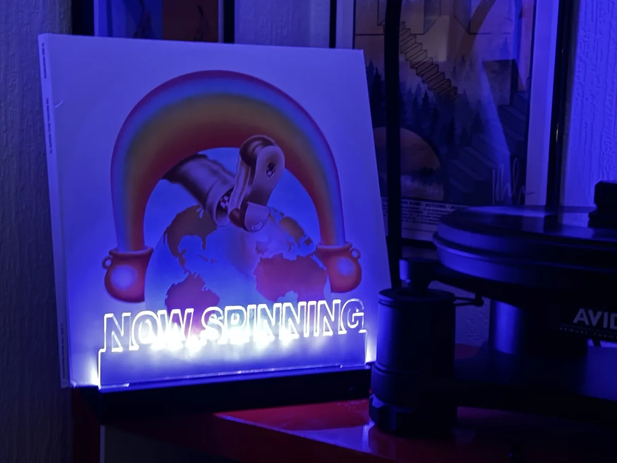 A glowing vinyl record sleeve display shows a rainbow arching over a globe with two pots at its ends, and the text 'NOW…