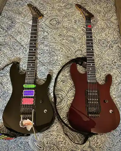 Two electric guitars lie side by side on a paisley-patterned fabric. The left guitar is black with colorful, mismatched…