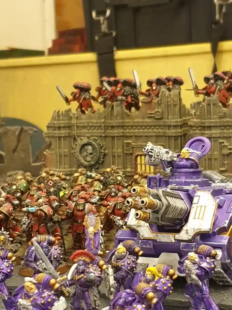 A photo of a Warhammer 40k tabletop battle scene shows purple-armored Space Marines in the foreground and red-armored…