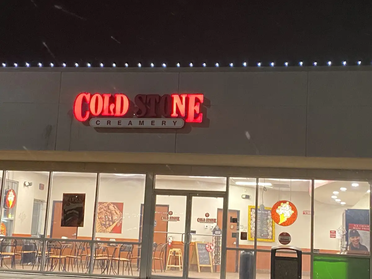 An exterior night photo of a Cold Stone Creamery storefront with its red neon sign illuminated above glass doors and windows…