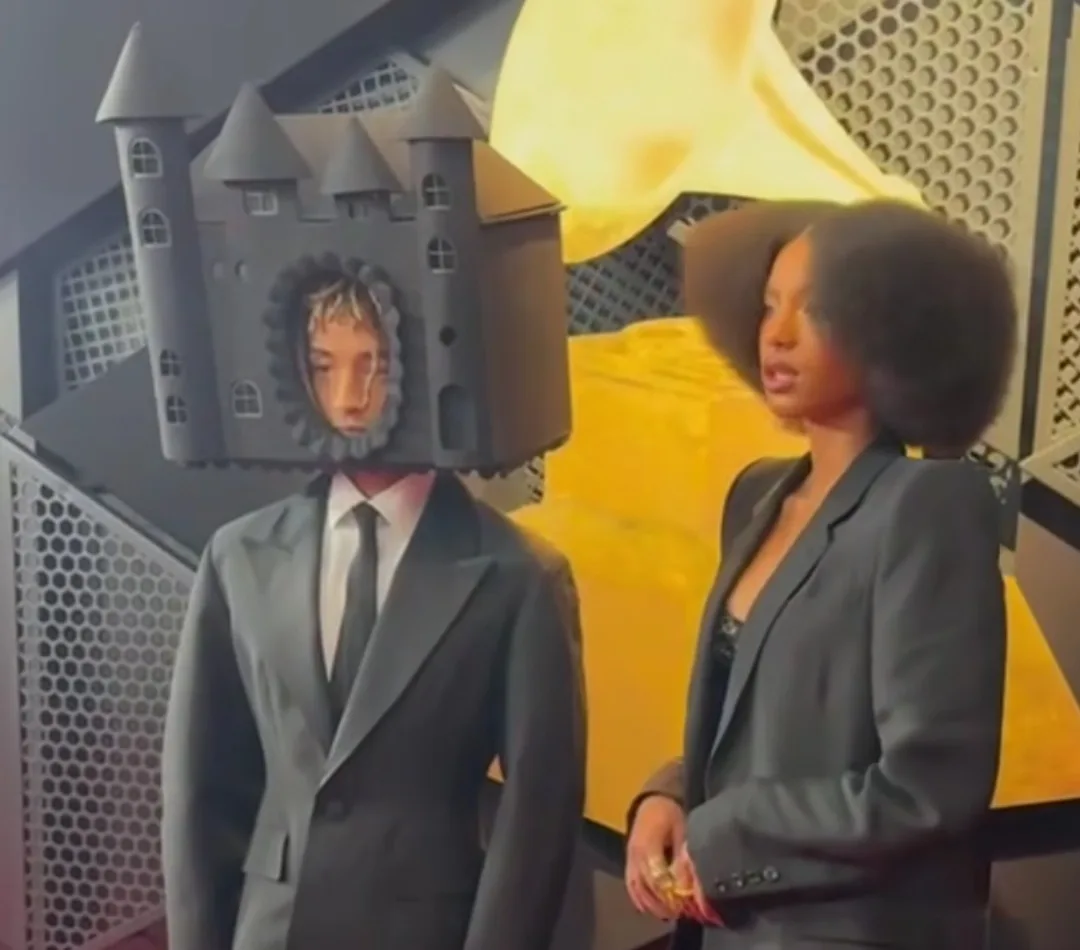 A person in a suit has a miniature castle model placed over their head, obscuring their face except for a small circular…