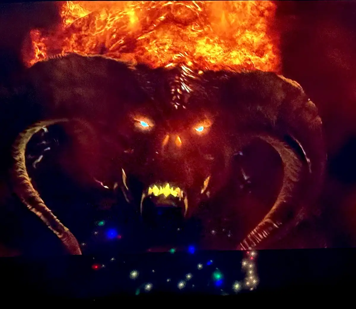 A menacing, horned demonic creature with glowing blue eyes and sharp teeth stares intensely, set against a fiery orange…