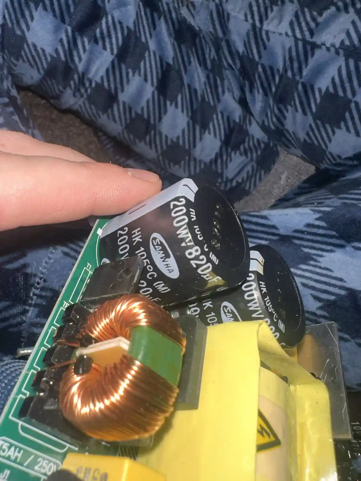 A close-up photo shows a person's finger holding a black cylindrical capacitor labeled '200MA 820uF' on a green circuit…