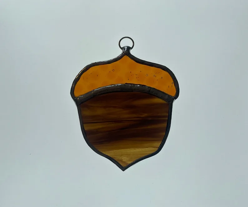 A stained glass ornament shaped like an acorn, featuring a bright amber cap and a darker amber body with wood grain texture…