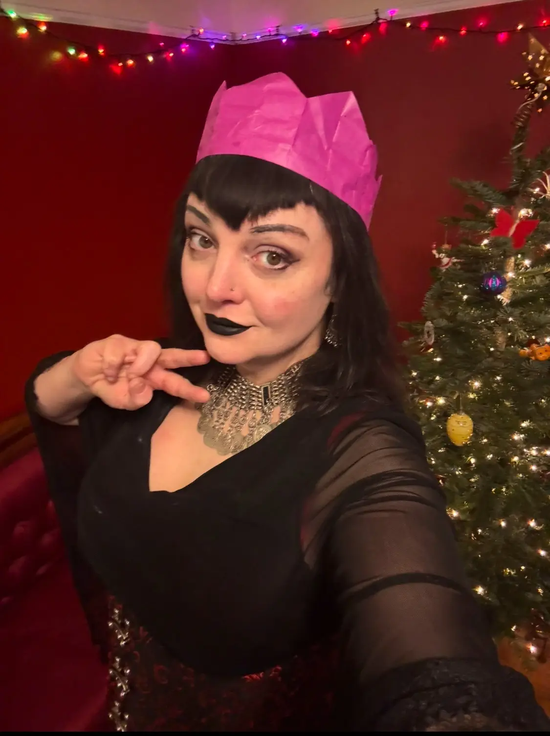 A woman with dark hair and black lipstick wears a bright pink paper crown and poses with a peace sign in front of a…