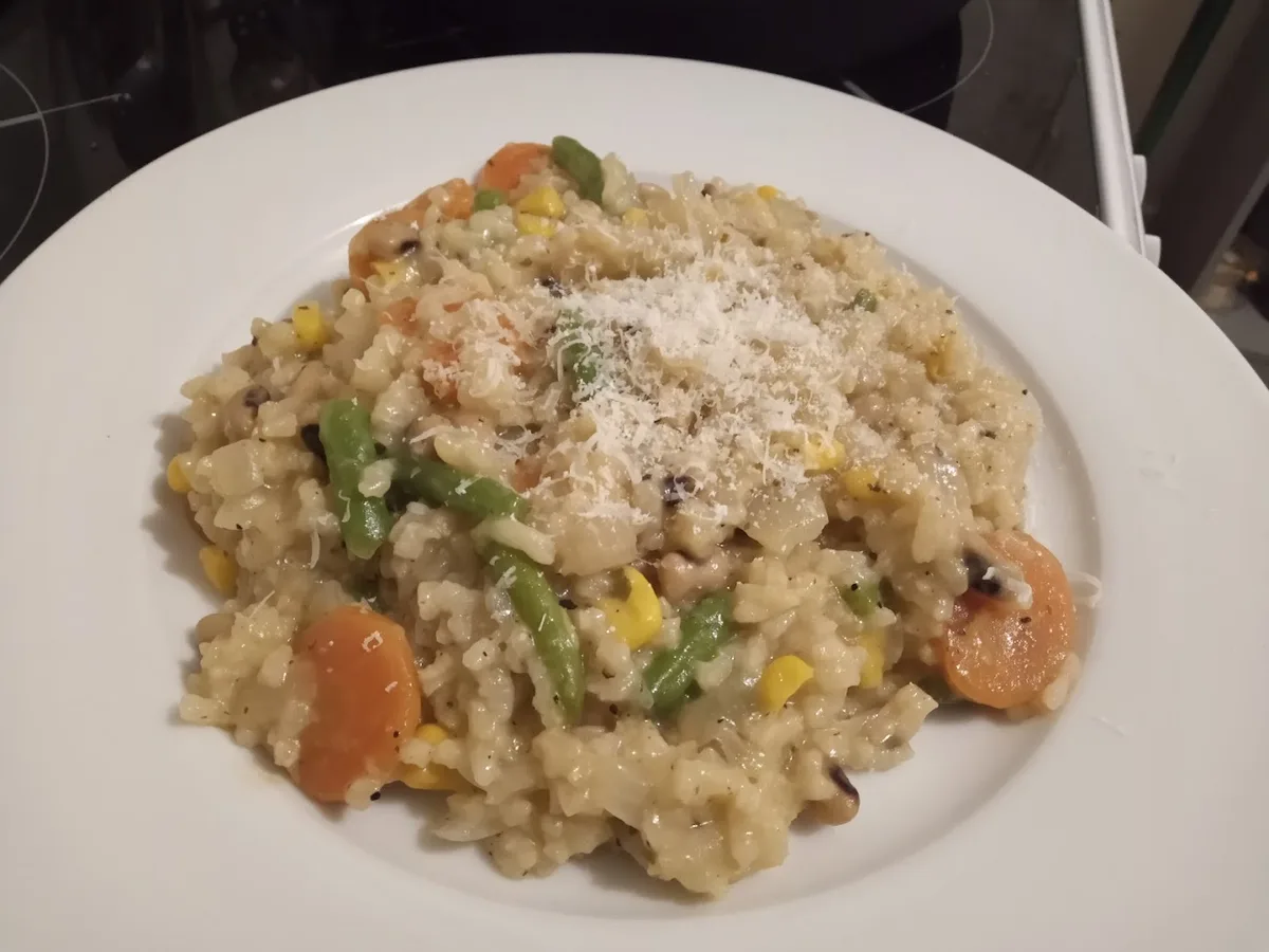 A close-up photo of a white plate holding a serving of risotto mixed with green beans, corn, and sliced carrots, topped with…