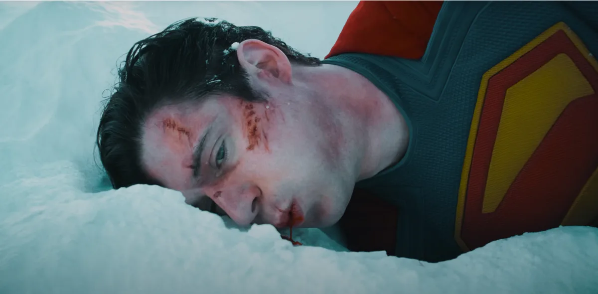 A close-up shot of a man in a Superman costume lying motionless in snow, with visible blood on his face and a trickle of…