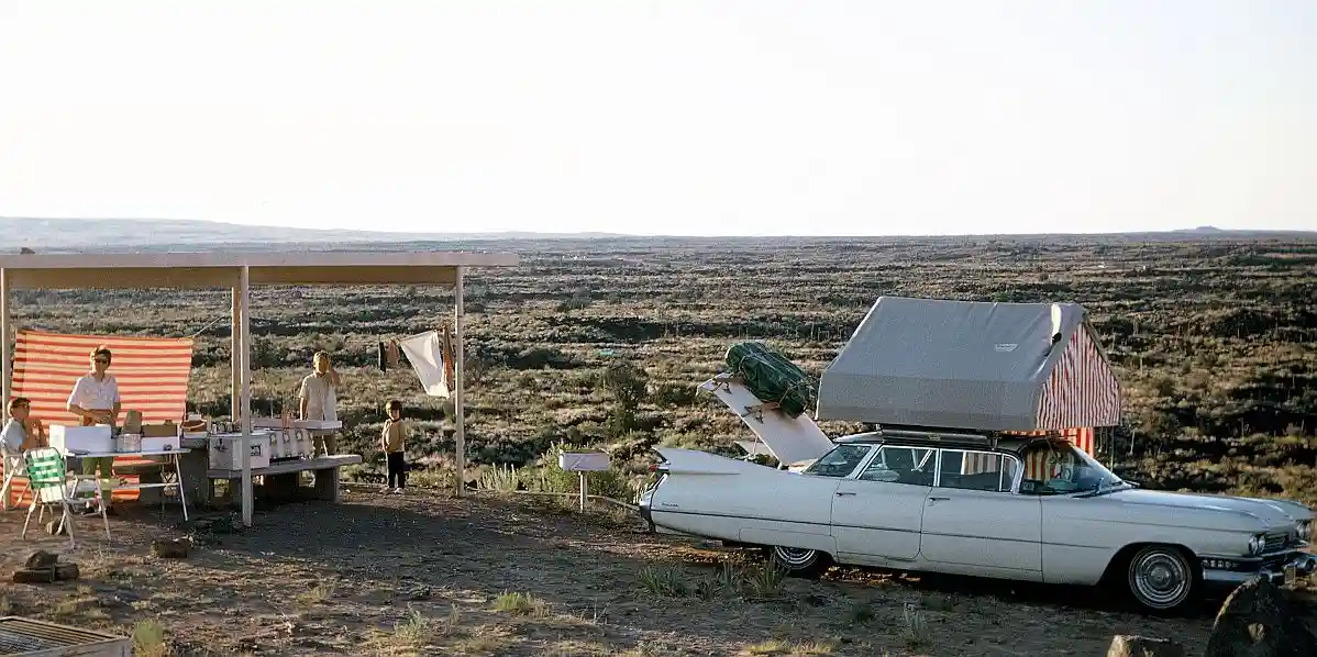 A vintage photo shows a family camping in a desert landscape with a white 1960s Cadillac with a rooftop tent parked nearby…