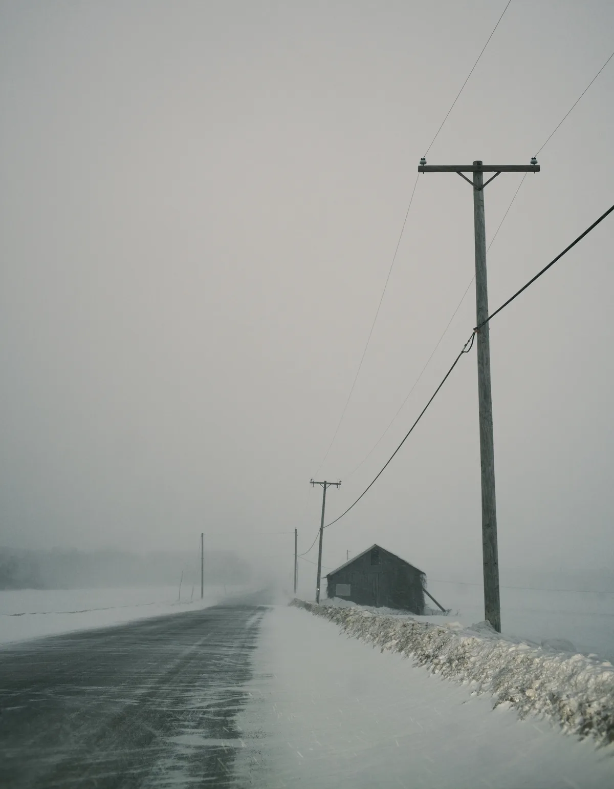 A desolate, snow-covered rural road stretches into a foggy, gray distance, flanked by wooden utility poles and a lone…