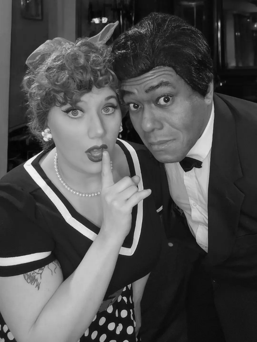 A black-and-white photo shows a woman in a vintage sailor-style dress with a polka-dot skirt, pearl necklace, and curled…