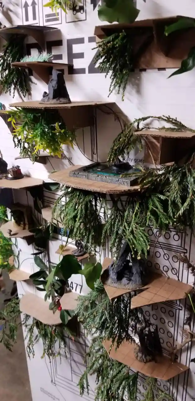 A wall-mounted display made of cardboard shelves, decorated with artificial greenery and miniature fantasy terrain pieces…