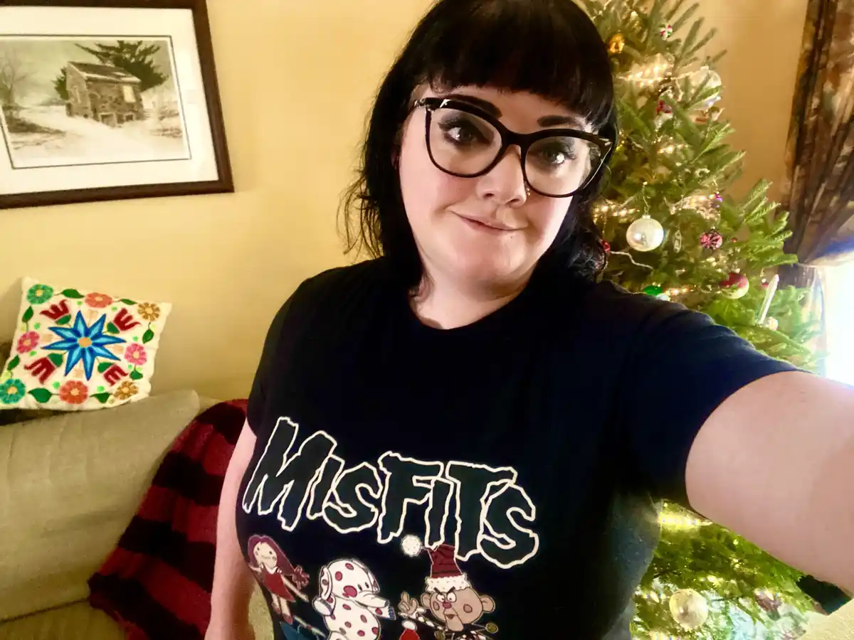A person with dark hair and glasses takes a selfie in front of a decorated christmas tree, wearing a black t-shirt with the…